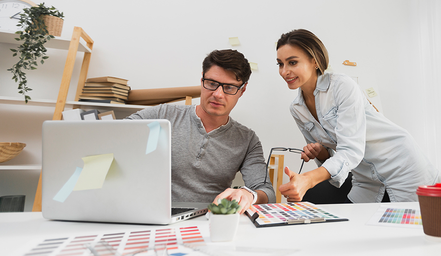 A man and a woman working together at a desk. The man is using a laptop, while the woman gestures with a thumbs-up. There are color swatches, papers, and a potted plant on the desk. Shelves with books and decor are in the background.