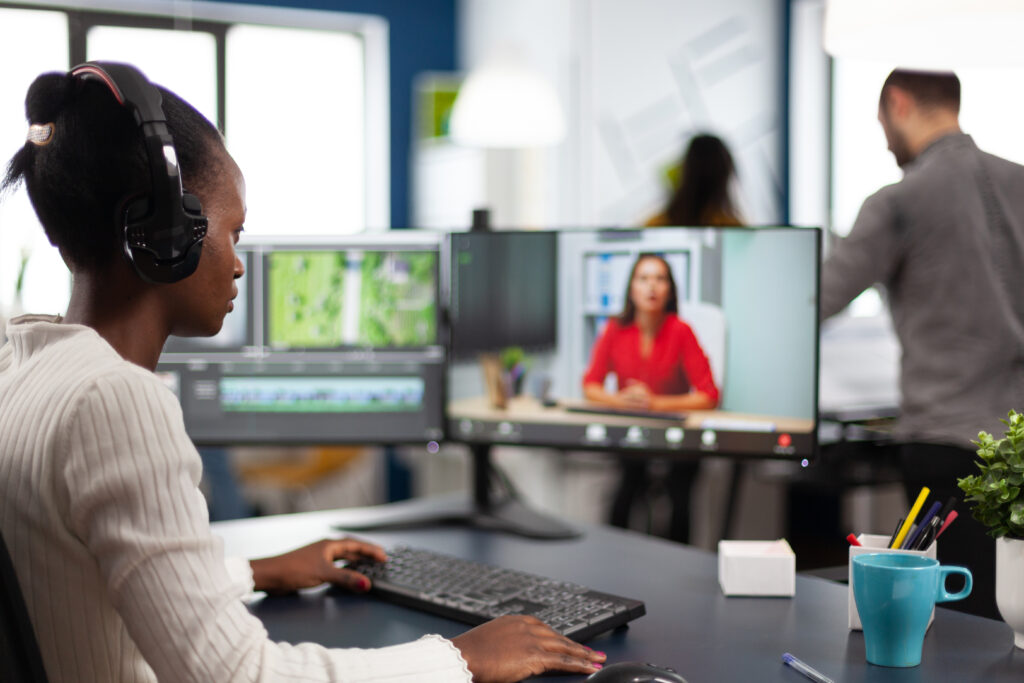 A person wearing headphones works at a remote IT support agency with dual monitors, perhaps providing IT support in Denver. One screen shows an editing timeline, while the other displays a video call with a person in a red shirt. Another person stands in the background. A mug and plant are on the desk.
