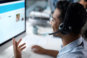 A person wearing a headset sits at a desk workin in a remote IT support services, smiling and gesturing as if engaged in an IT support call. The scene suggests they're part of an IT support team in Denver, with blurred office elements visible in the background.